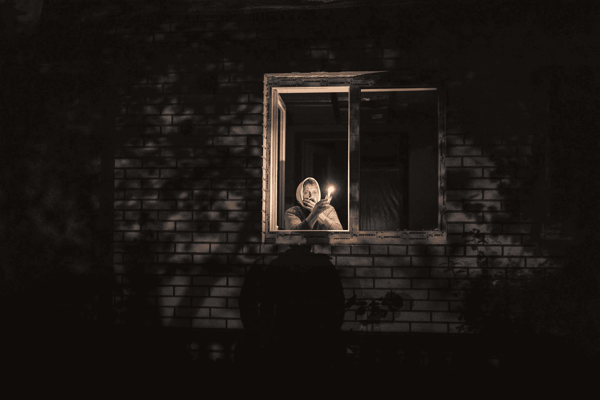 A person holding a small flame in a dark window of a brick building, reflecting the sombre reality of the Ukraine war.