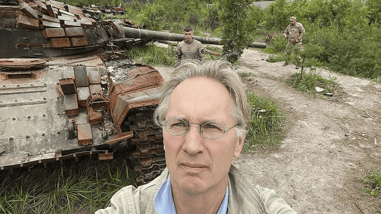 Jaap Scholten selfie in front of a destroyed tank with military personnel in the background, during the Ukraine war.