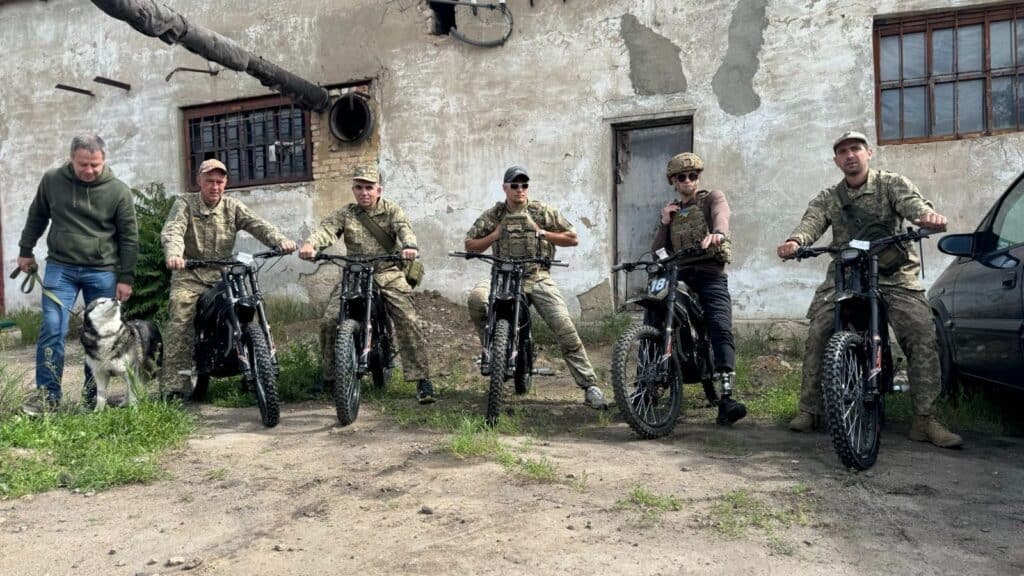 Four Ukrainian soldiers on donated Protect Ukraine electric motorcycles, with a man and a dog standing alongside them.