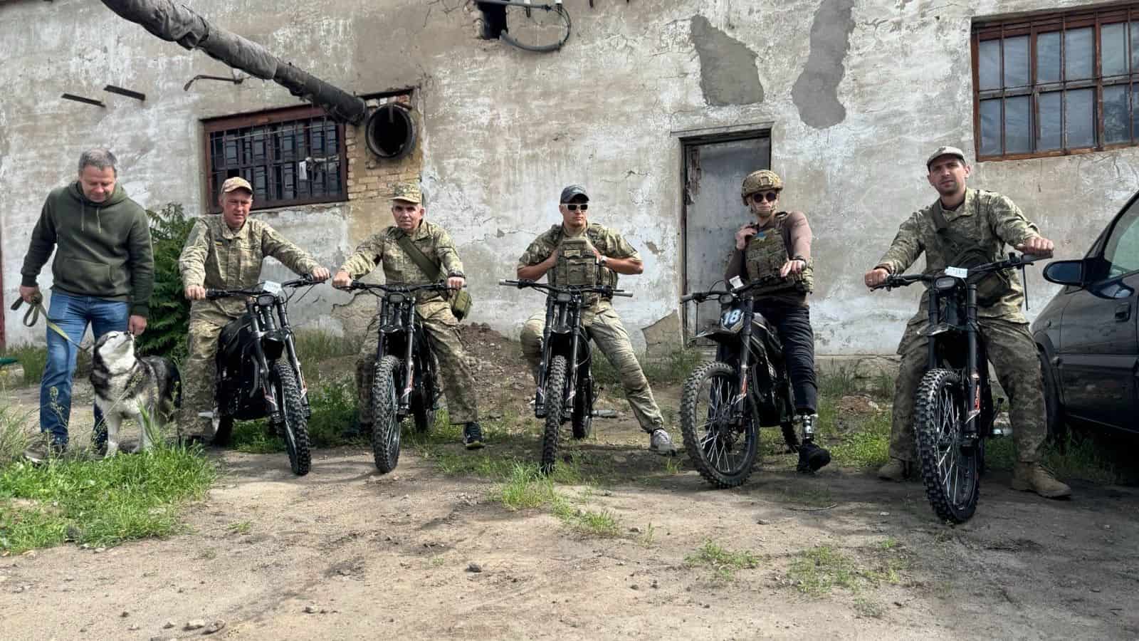Four Ukrainian soldiers on donated Protect Ukraine electric motorcycles, with a man and a dog standing alongside them.